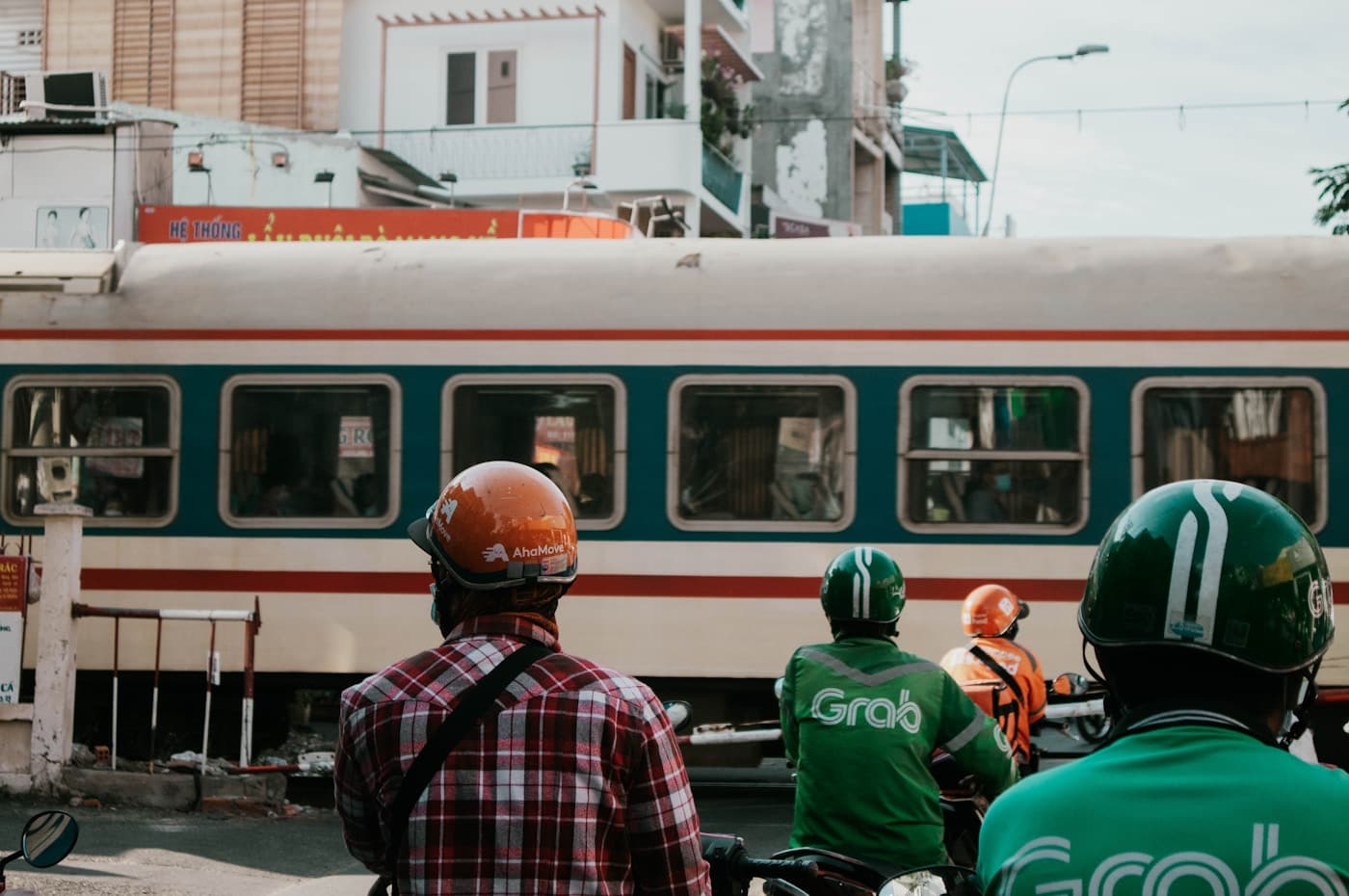 Пассажирский поезд на железнодорожном переезде, перед ним ждут водители Grab в зелёных шлемах