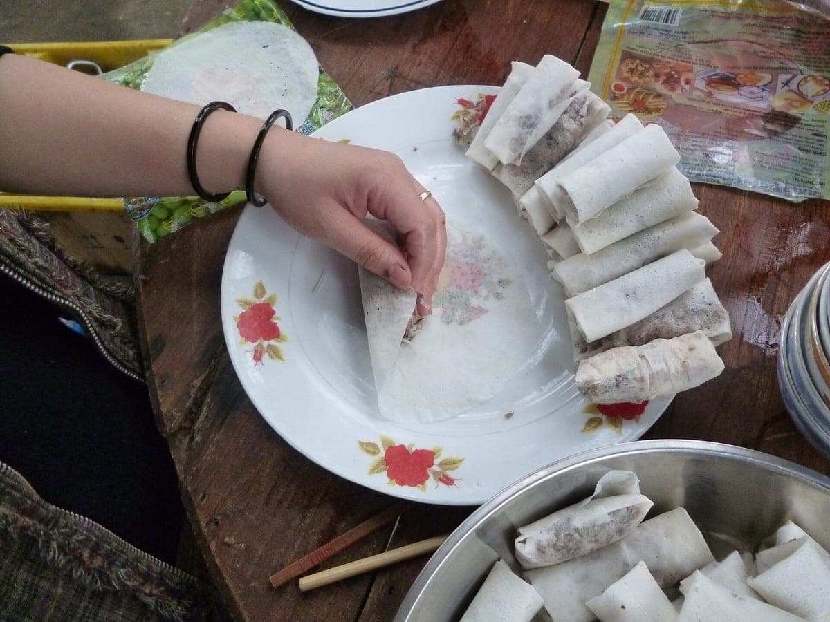 Рука заворачивает начинку в лист рисовой бумаги на тарелке, рядом ряд готовых сырых нэмов